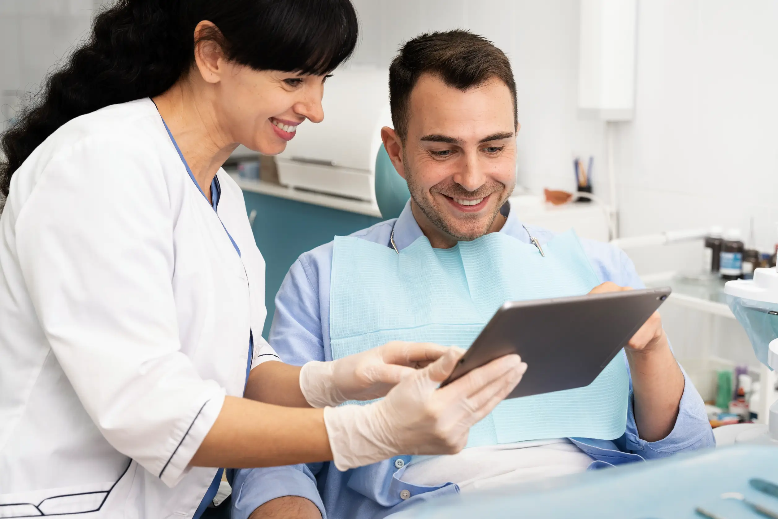 A female dentist in a white coat and gloves is pointing at a tablet she is holding, showing it to a male patient sitting in a dental chair